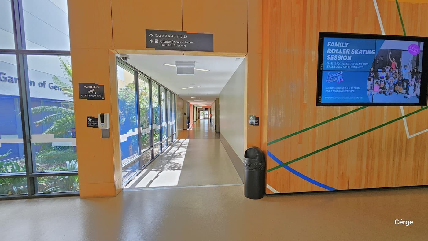 Corridor after passing through the turnstiles that regulate entry and right next to the wall that imitates the court floor with green, white, and yellow lines with a digital monitor mounted on it. On the top wall of the corridor opening is a black sign containing the directions for Courts 3 & 4, 9 to 12, Change Rooms, Toilets, First Aids, and Lockers. Also found here is a sanitizing dispenser and a CCTV sign on the left wall and a black garbage bin on the right wall.