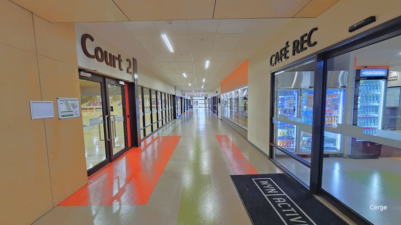 Café REC at Eagle Stadium is directly opposite the Court 2 entrance doors. Court 2 has manually operated glass doors with stainless pull handles while Café REC has automatic, glass sliding doors. There is a black mat with WYN and ACTIVE in front of the Café entrance doors. Both amenities have glass windows with black metal frames. The floor, wall, and door have orange-painted sections. Seen through the Café glass door is the fridge containing different cold drinks.