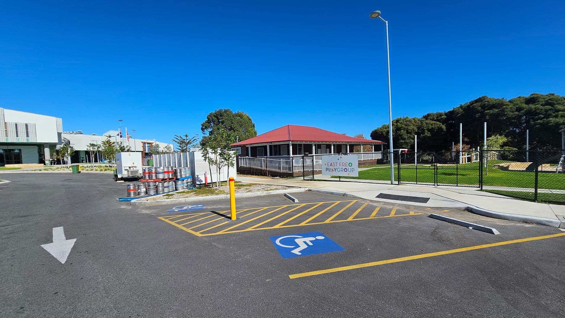 A parking area with a marked disabled parking space in the foreground. To the left, there are several metal barrels stacked near a building with a modern design. In the centre, a building with a red roof and a sign reading "East Freo Playgroup" is visible, surrounded by a white fence. To the right, a grassy area with trees and a fenced playground. A tall streetlamp stands near the playgroup building. The sky is clear and blue.