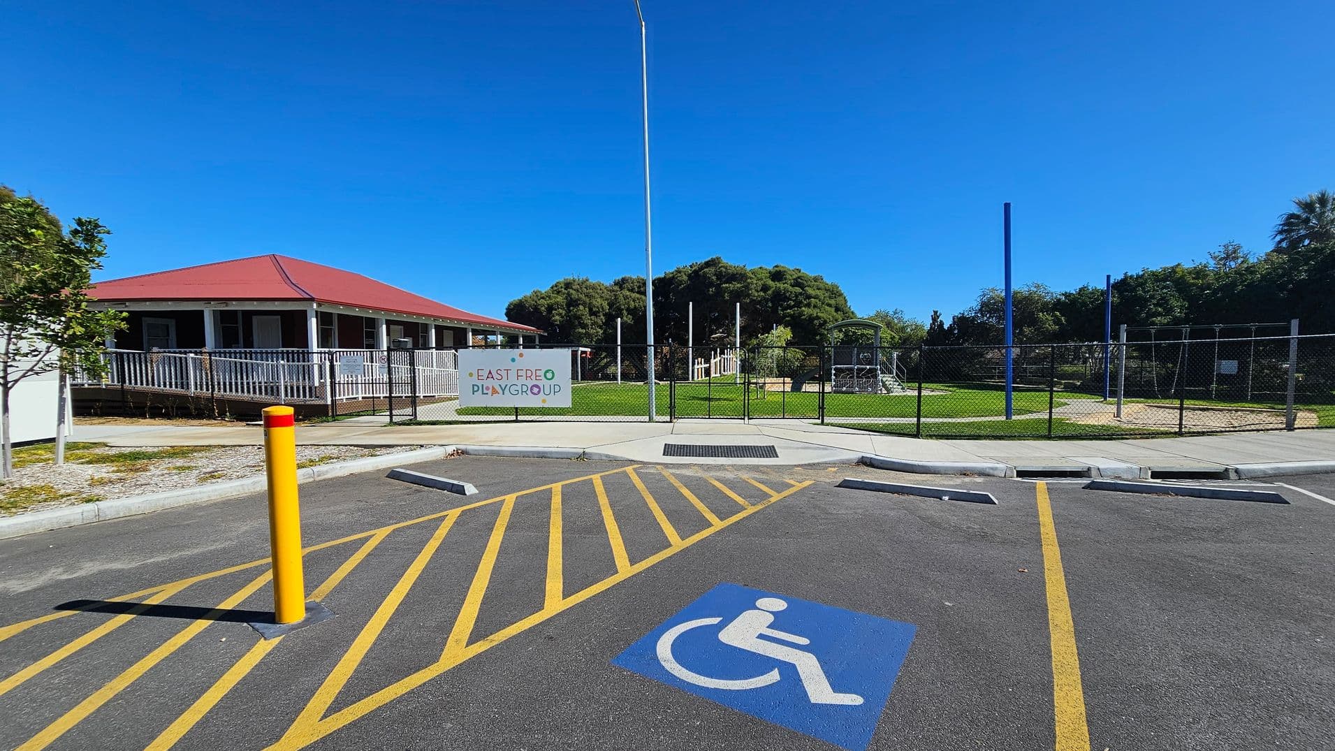 A parking area with a marked disabled parking space in the foreground, featuring a blue wheelchair symbol. Yellow lines and a yellow bollard are visible. In the background, a building with a red roof and a white railing is on the left. A sign reading "East Freo Playgroup" is attached to a fence, with a grassy area and playground equipment behind it. Trees are visible in the distance.