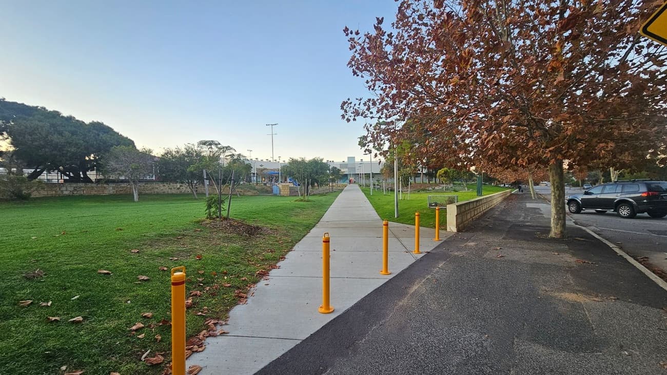 A pathway extends into the distance, flanked by green grass on the left and a row of trees with brown leaves on the right. Yellow bollards line the path's entrance. To the right, a parked car is visible on the road. In the background, there are more trees and a building with a playground. The sky is clear, and fallen leaves are scattered on the ground.