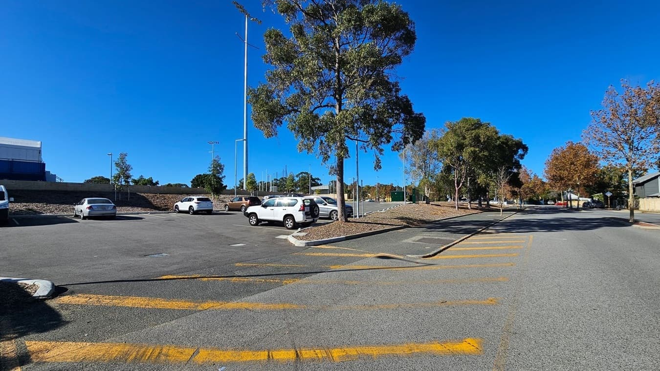 A parking area with several cars is on the left, bordered by a low stone wall and a few small trees. A tall tree stands in the centre, with a street extending to the right, lined with more trees showing autumn foliage. The ground is paved with yellow markings, and tall floodlights are visible in the background. The sky is clear and blue.
