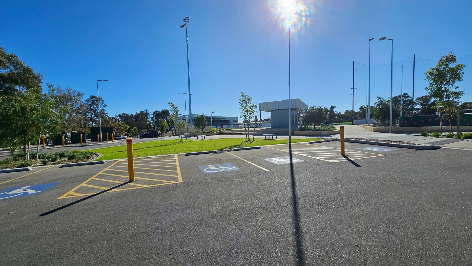 A parking area with marked disabled spaces in the foreground, featuring yellow and blue markings. In the middle, there is a grassy area with small trees and benches. The background shows a building with a sloped roof and several tall light poles. Trees and additional parking spaces are visible on the left and right sides. The sky is clear and sunny, casting shadows on the ground.