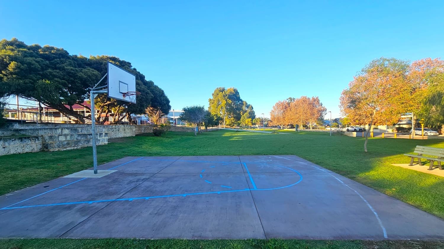 A small outdoor basketball court with a single hoop on the left. The court has blue lines and is surrounded by grass. In the background, there are several trees with green and brown leaves. To the right, a wooden bench is visible. A stone wall runs along the left side, and there are buildings and parked cars in the distance. The sky is clear and blue.