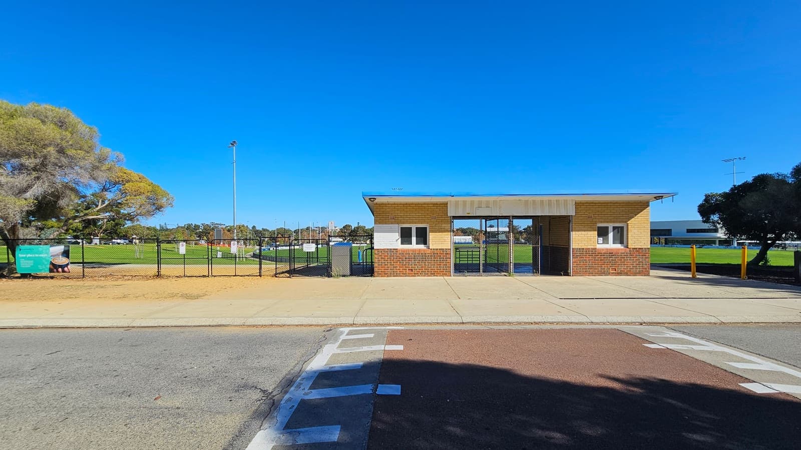 A small brick building with a metal roof stands in the centre, featuring two windows. To the left, a fence with a sign encloses a grassy field, with a tree nearby. On the right, more grass and a few trees are visible. The foreground has a paved walkway and a road with painted lines. The sky is clear and blue.