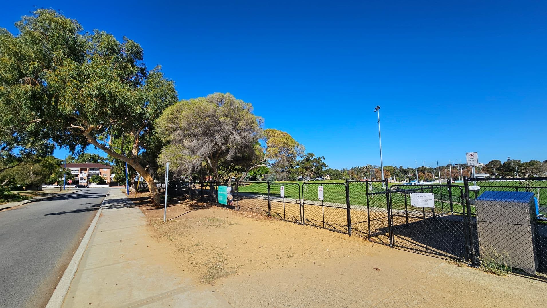 A paved footpath runs alongside a road on the left, bordered by large trees providing shade. On the right, a fenced sports field with green grass is visible, featuring tall floodlights. Signs are attached to the fence, and a small structure is near the entrance. In the background, there are more trees and a few buildings. The sky is clear and blue.