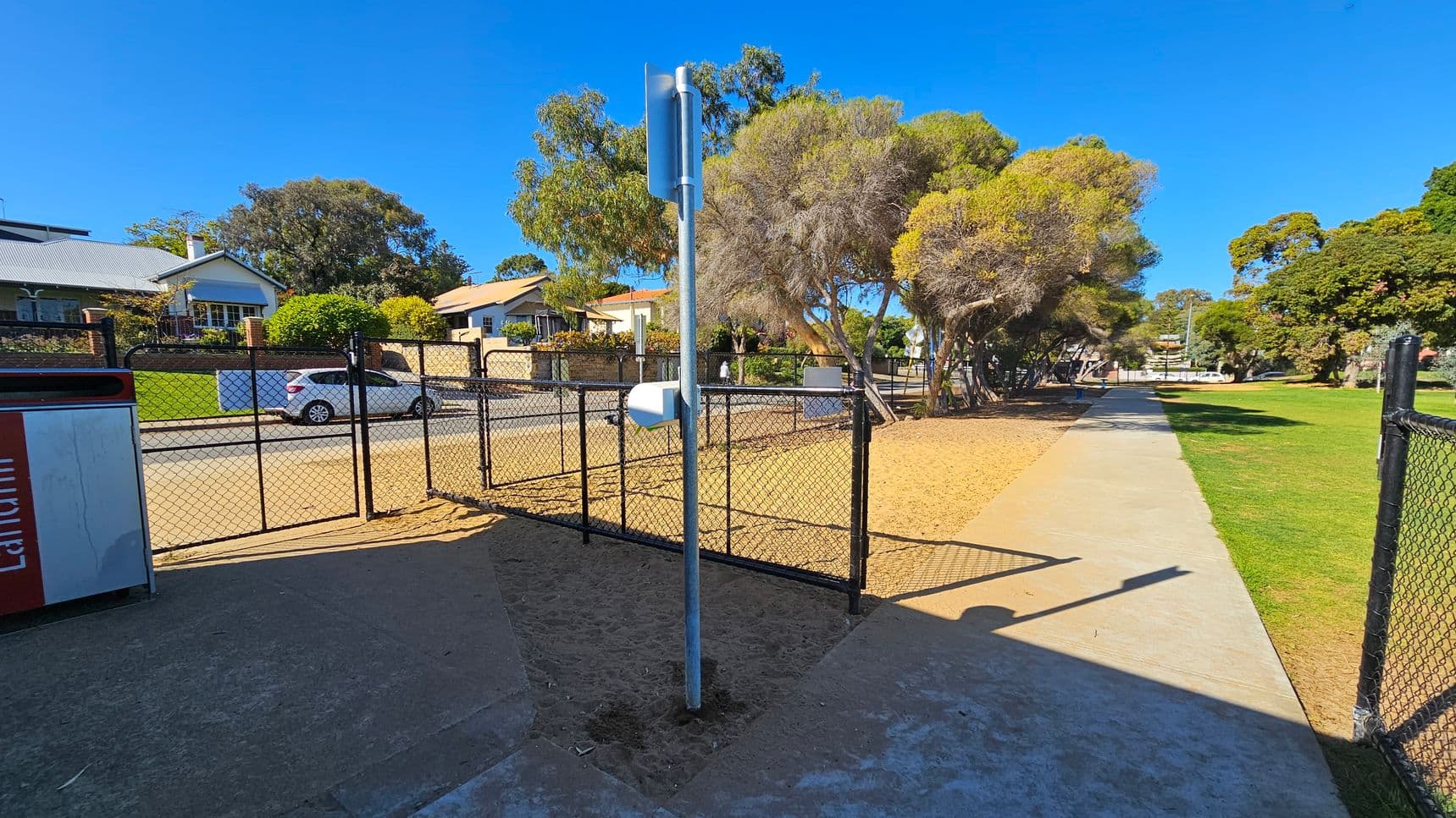 A paved path runs through a park, bordered by a black chain-link fence. In the centre, a metal pole with attached equipment stands on a sandy patch. To the left, a bin is visible. Beyond the fence, a road with a white car and houses with well-kept gardens can be seen. On the right, the path continues alongside a grassy area with trees. The sky is clear and blue.