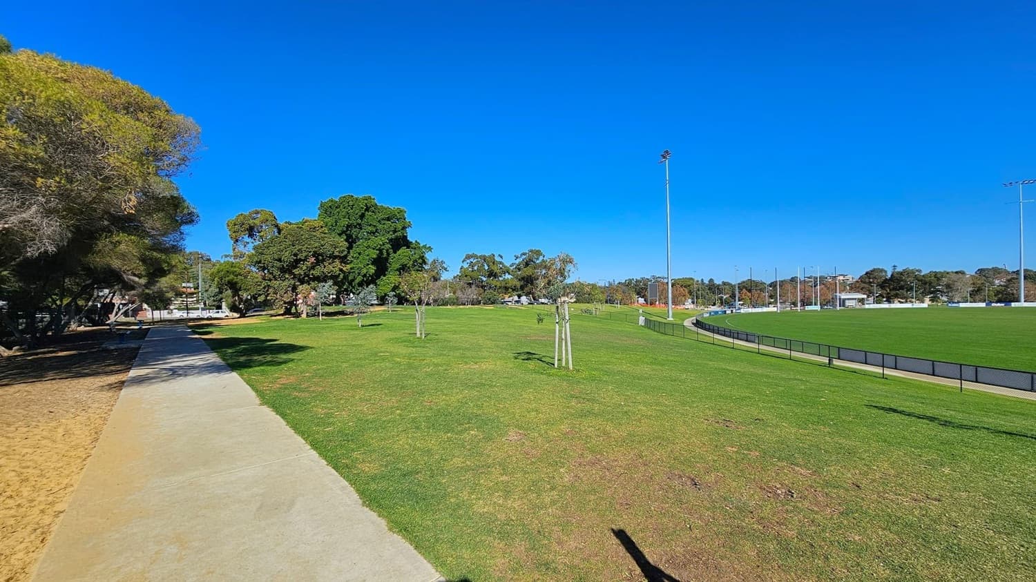 A wide grassy field with a concrete path on the left, bordered by trees providing shade. On the right, a fenced sports field with tall floodlights. The background features more trees and some buildings. The ground is mostly grass with patches of dirt near the path.