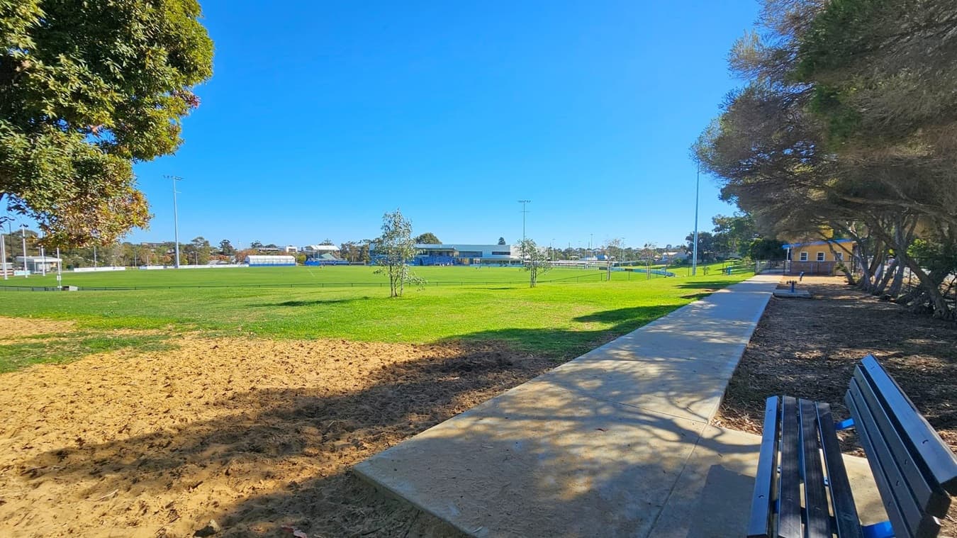 A park scene with a large grassy field in the centre, bordered by a paved path on the right. Benches are placed along the path, with trees providing shade. On the left, a large tree casts shadows on the ground. In the background, there are buildings and sports facilities, including floodlights. The sky is clear and blue.