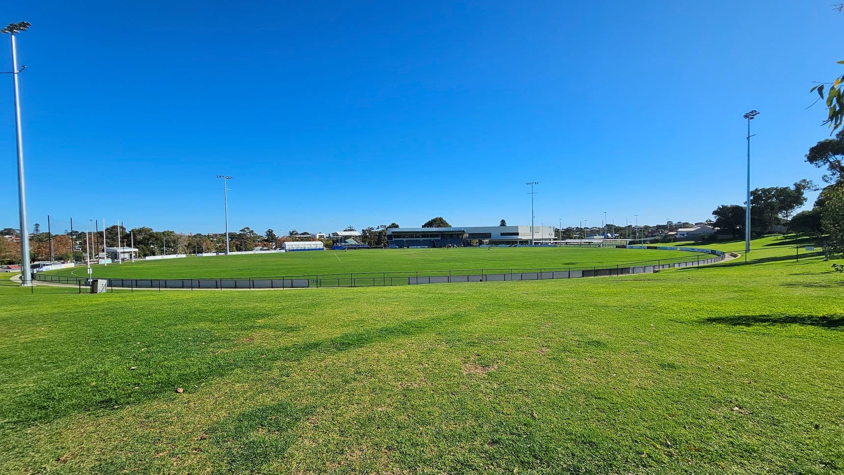 A large, open sports field with a well-maintained grassy surface. Surrounding the field is a low metal fence. Tall floodlights are positioned around the perimeter. In the background, there are several buildings, including a large structure with a flat roof. Trees line the horizon, and a clear blue sky is overhead. The foreground features a gently sloping grassy area.