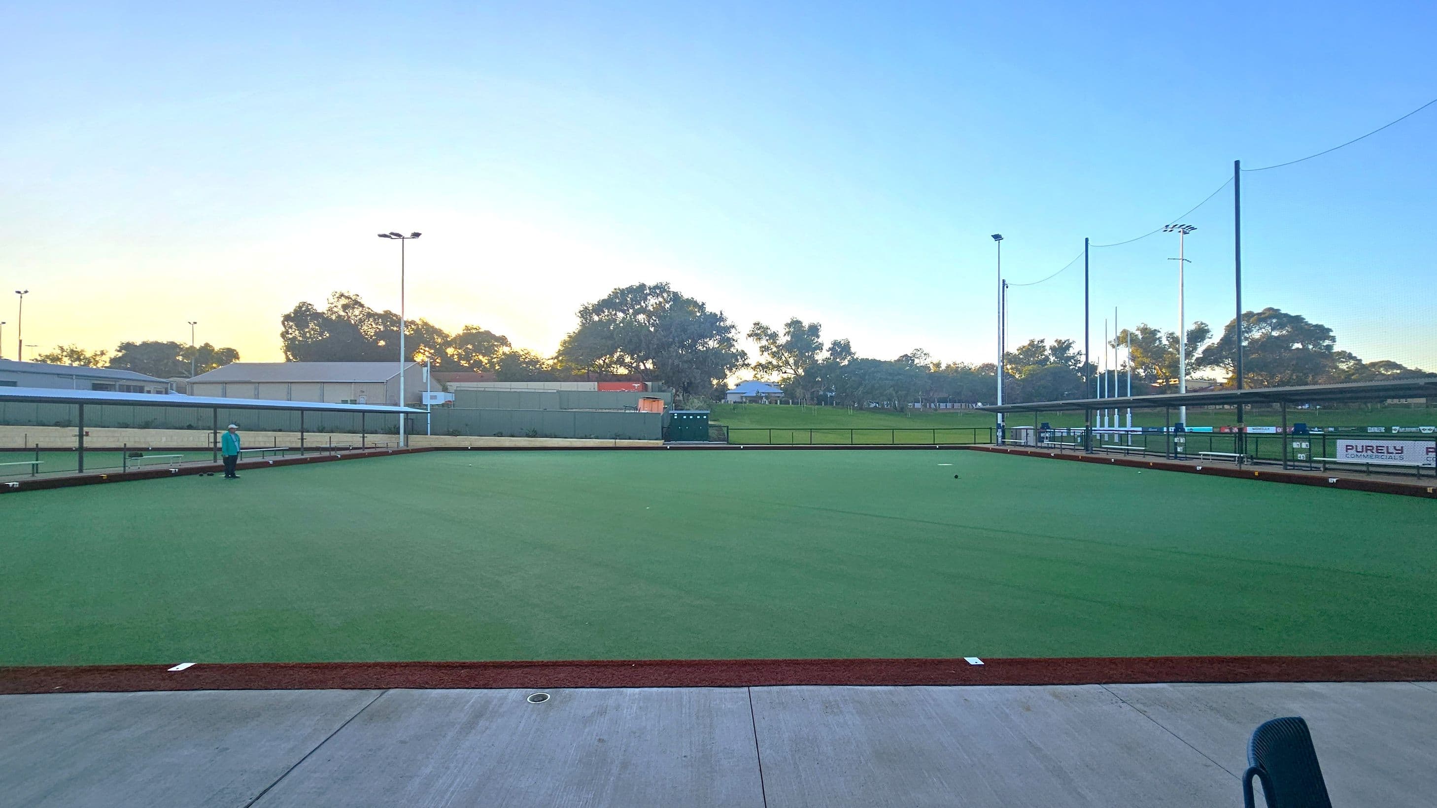 A lawn bowling green with a well-maintained grass surface is in the centre. A person stands on the left side near the edge of the green. Surrounding the green are low barriers and several tall floodlights. In the background, there are trees and a few buildings. The sky is clear with a hint of sunset or sunrise. The foreground features a concrete area with a chair partially visible.