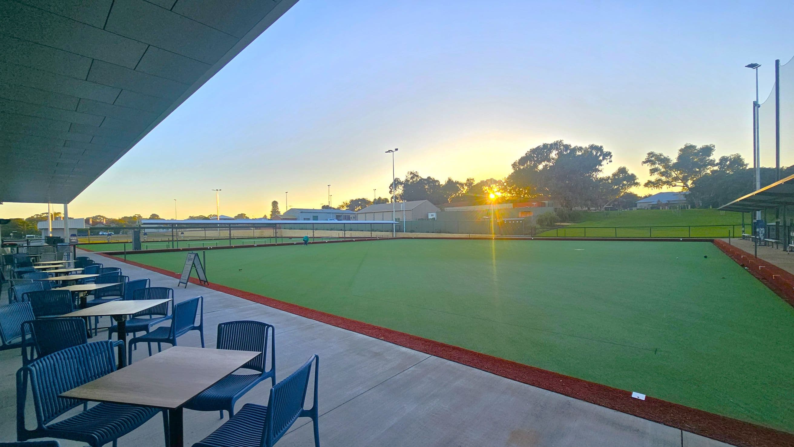 On the left, there are several tables with blue chairs arranged on a concrete patio under a slanted roof. The middle features a large, well-maintained green lawn, possibly for sports, bordered by a red pathway. In the background, the sun is setting behind a row of trees and buildings, casting a warm glow. The right side shows more of the lawn and a continuation of the pathway, with additional seating under a smaller roof.