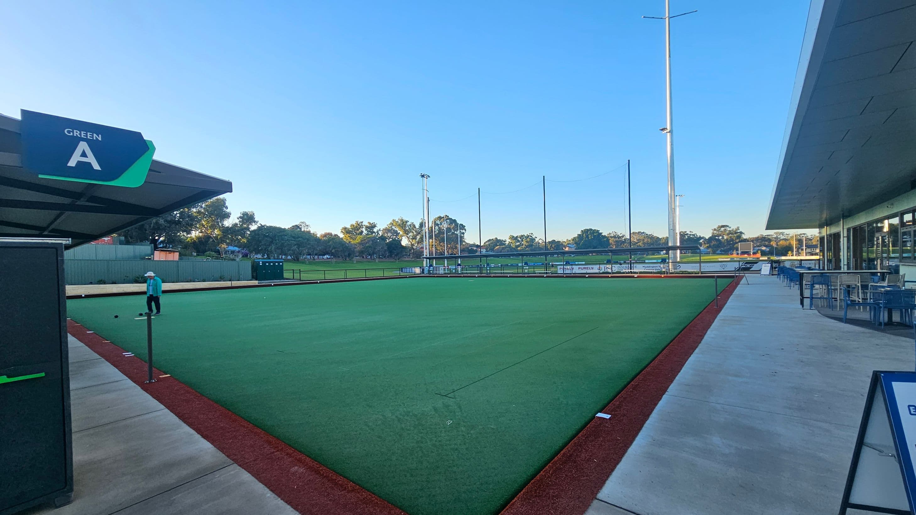 A lawn bowling green with a sign labelled "Green A" on the left. A person in a blue shirt and hat is playing on the green. The foreground has a red border around the green. On the right, there is a modern building with large windows and outdoor seating with tables and chairs. Tall light poles are visible in the background, along with trees and a fence. The sky is clear.