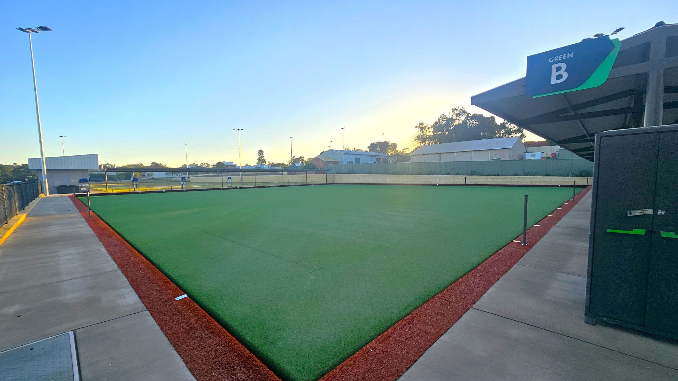 A well-maintained lawn bowling green is in the centre, bordered by a red edge. On the right, a structure with a sign reading "Green B" is visible. To the left, a concrete pathway runs alongside a fence, with a tall light pole nearby. In the background, there are several buildings and trees, with a clear sky above.
