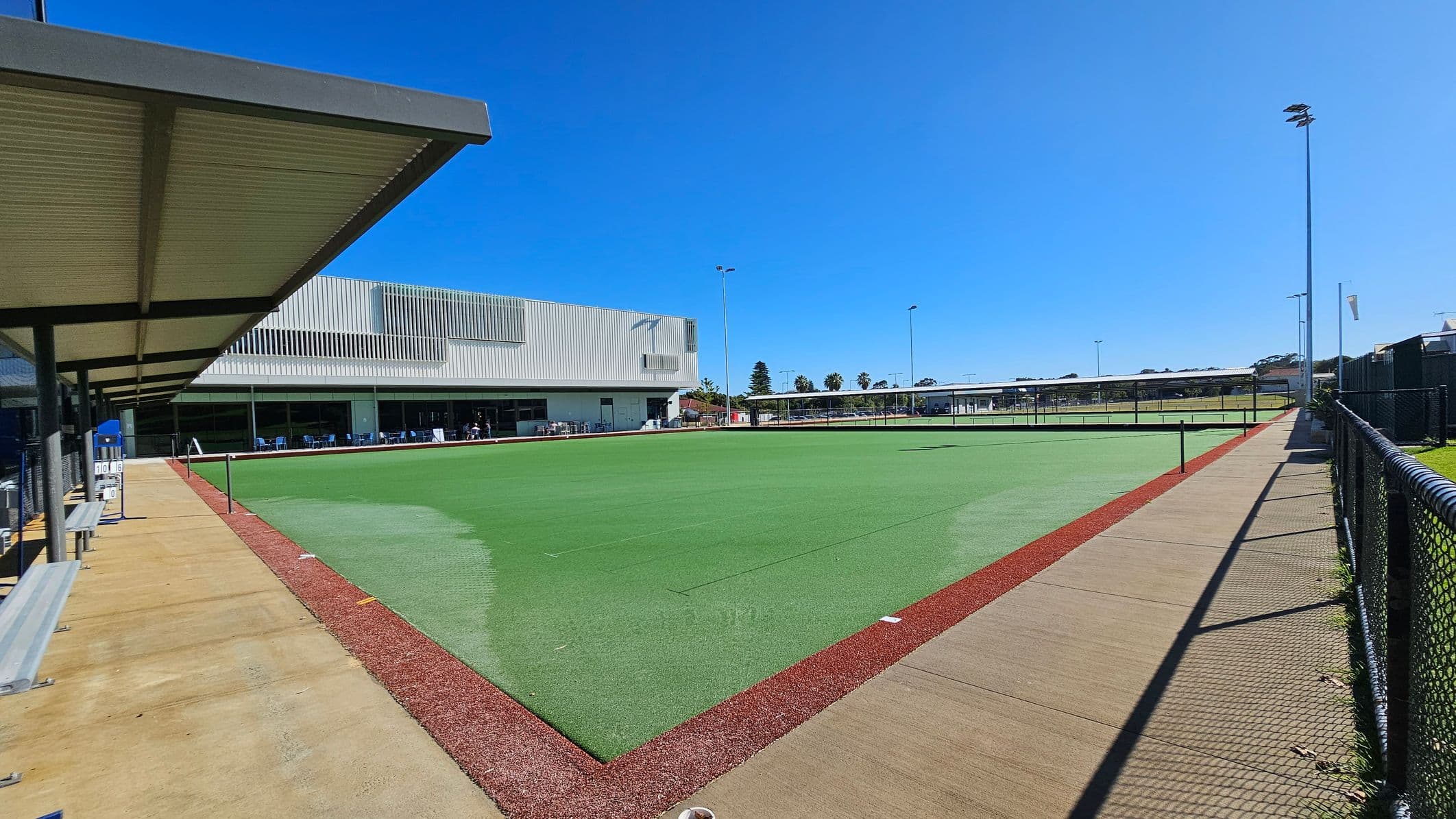 A sports facility with a large green artificial turf field in the centre, bordered by a red track. On the left, a covered walkway with benches and a vending machine. The building in the background has a modern design with large windows and a corrugated metal facade. On the right, a chain-link fence runs alongside a concrete path. Tall floodlights are positioned around the field, and there are trees and additional structures in the distance.