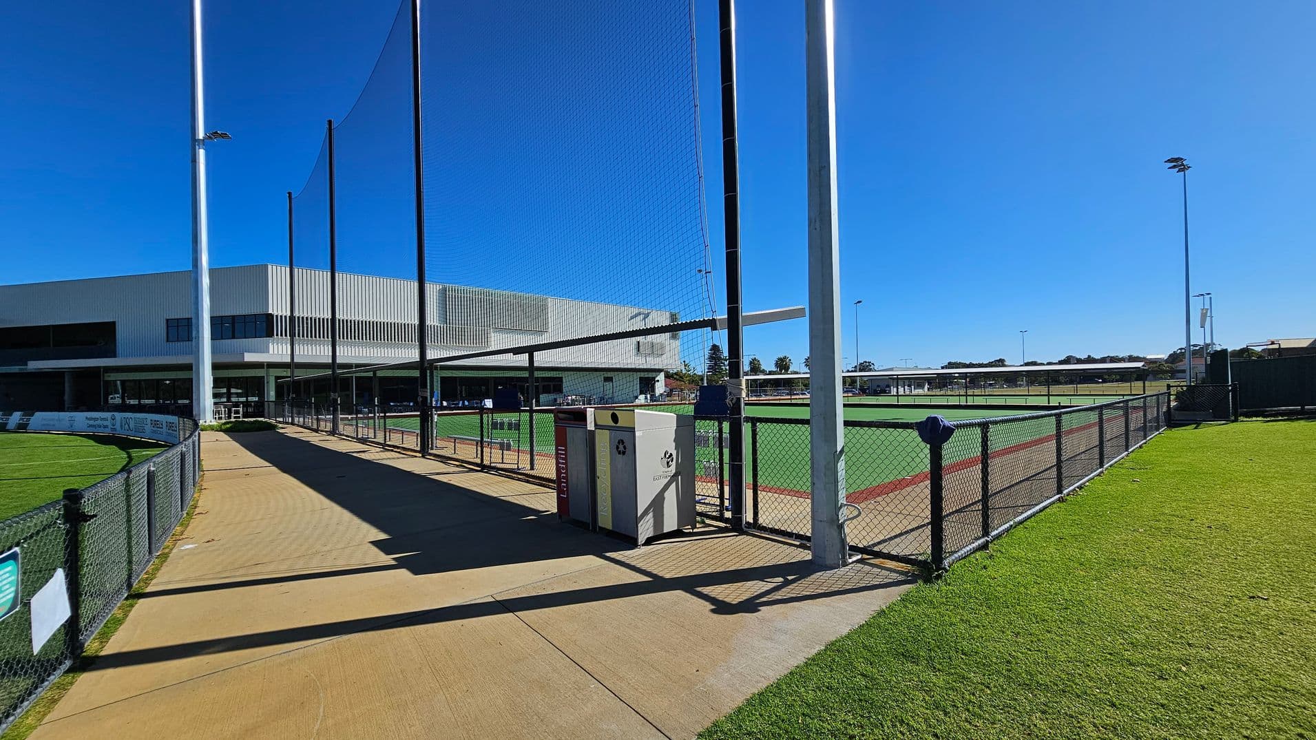 A sports facility with a modern building on the left, featuring large windows and a flat roof. In the centre, a pathway runs alongside a chain-link fence with a net above it. Two bins are positioned near the fence. On the right, a green sports field is visible, bordered by more fencing. Tall floodlights are scattered around the area. The ground is a mix of concrete and grass.