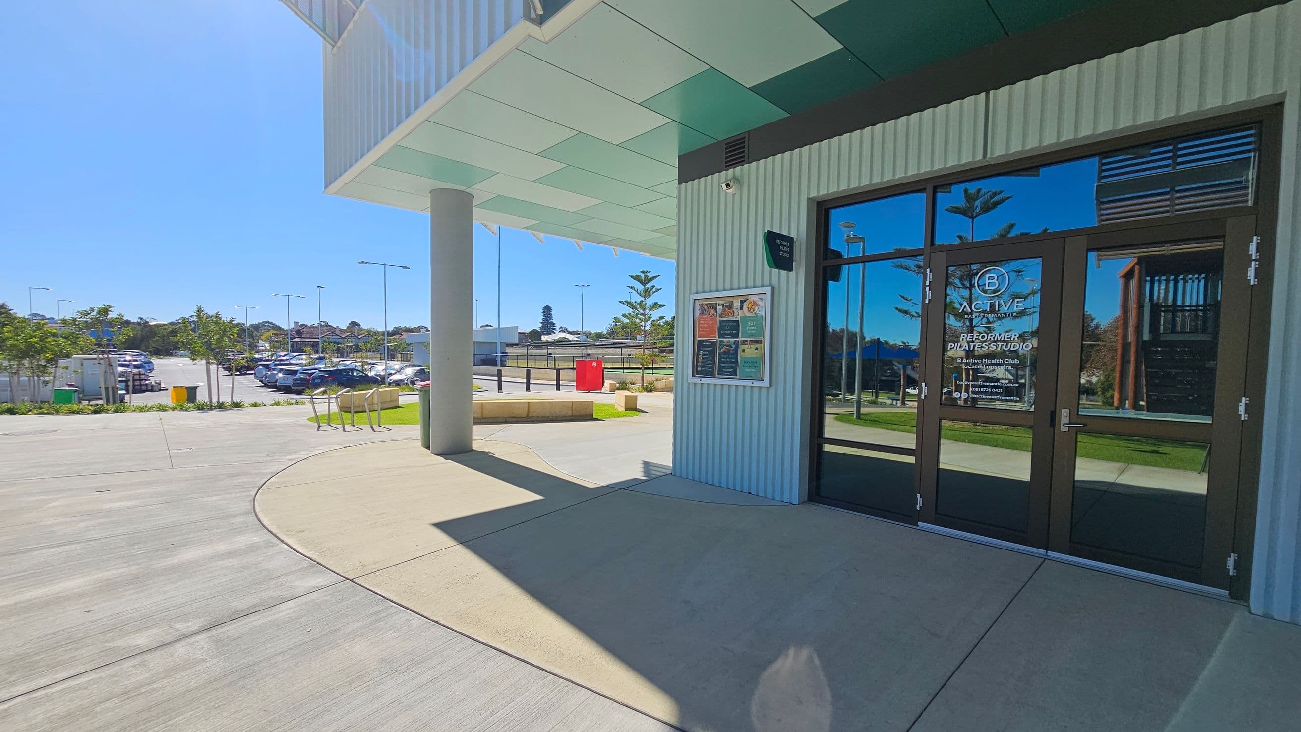 A modern building entrance with glass doors on the right, displaying signage for a Pilates studio. The exterior wall is corrugated metal with a notice board. The ceiling has a geometric design. In the background, a car park is visible with several parked cars and small trees lining the area. A red bin and a few streetlights are also present. The ground is a mix of concrete and grass, with a curved pathway leading towards the entrance.