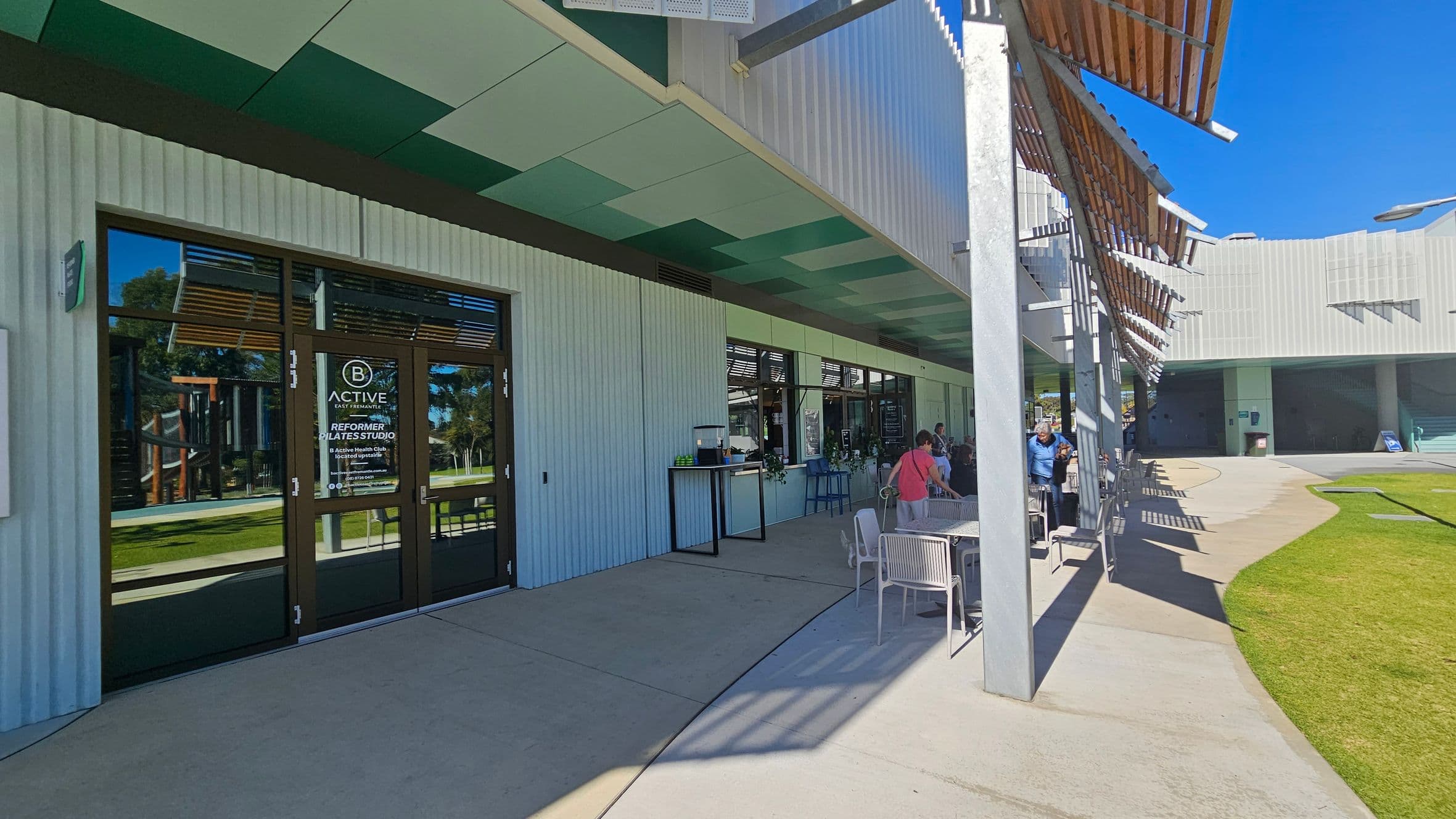 A modern building with a corrugated exterior and a shaded outdoor seating area. On the left, there are glass doors with signage for a Pilates studio. The ceiling above is patterned with green and white panels. To the right, people are seated at tables under a pergola with wooden slats. The ground is a mix of concrete and grass, with a curved pathway leading towards the background.
