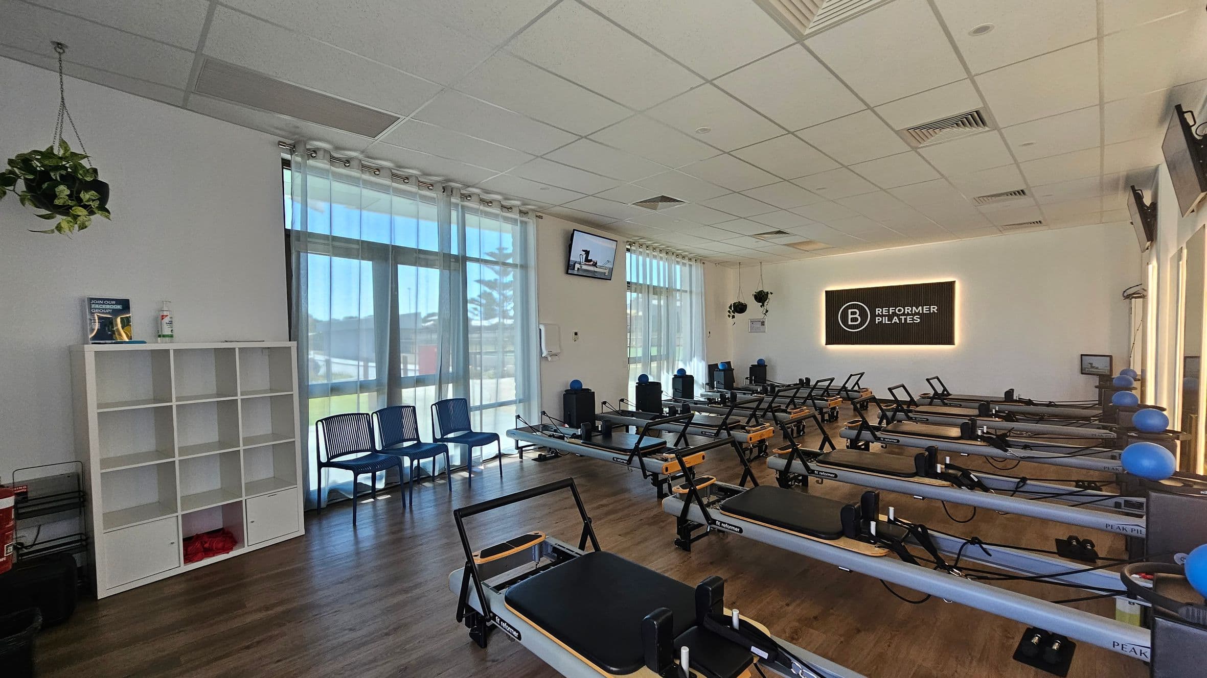 A Pilates studio with a wooden floor and a white ceiling. On the left, there is a white shelving unit with a book and sanitiser on top, and a hanging plant above. Blue chairs are lined up against the wall with large windows covered by sheer curtains. In the centre, several Pilates reformer machines are arranged in rows. On the right, a sign reads "Reformer Pilates" on the wall. Blue exercise balls are placed on some machines. Televisions are mounted on the walls.
