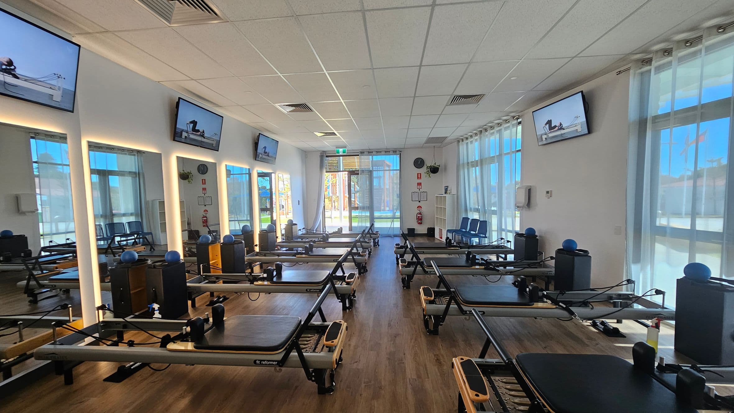 A spacious Pilates studio with multiple reformer machines arranged in rows on a wooden floor. The left wall features large mirrors with illuminated frames and mounted screens displaying exercise videos. The right side has large windows with sheer curtains, allowing natural light to enter. At the far end, there's a glass door leading outside, with a clock and safety equipment on the wall. The ceiling is fitted with white tiles and recessed lighting.