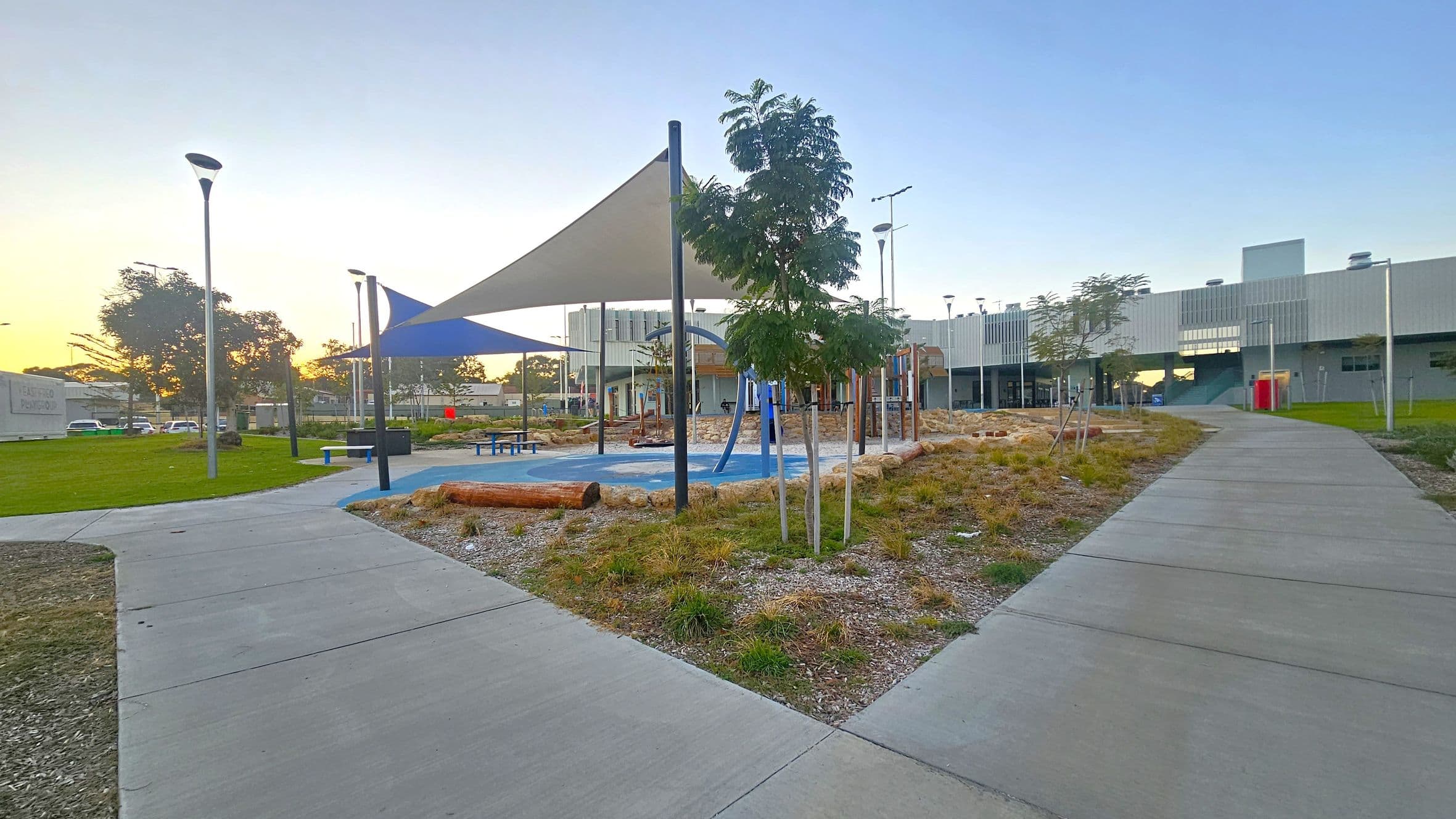 A playground area with a blue and white shade sail in the centre, surrounded by a mix of grass and gravel. There are small trees and wooden logs scattered around. On the left, a grassy area with a few cars and a sign. On the right, a modern building with large windows and a pathway leading towards it. The ground is paved with concrete paths, and several street lamps are visible throughout the scene.