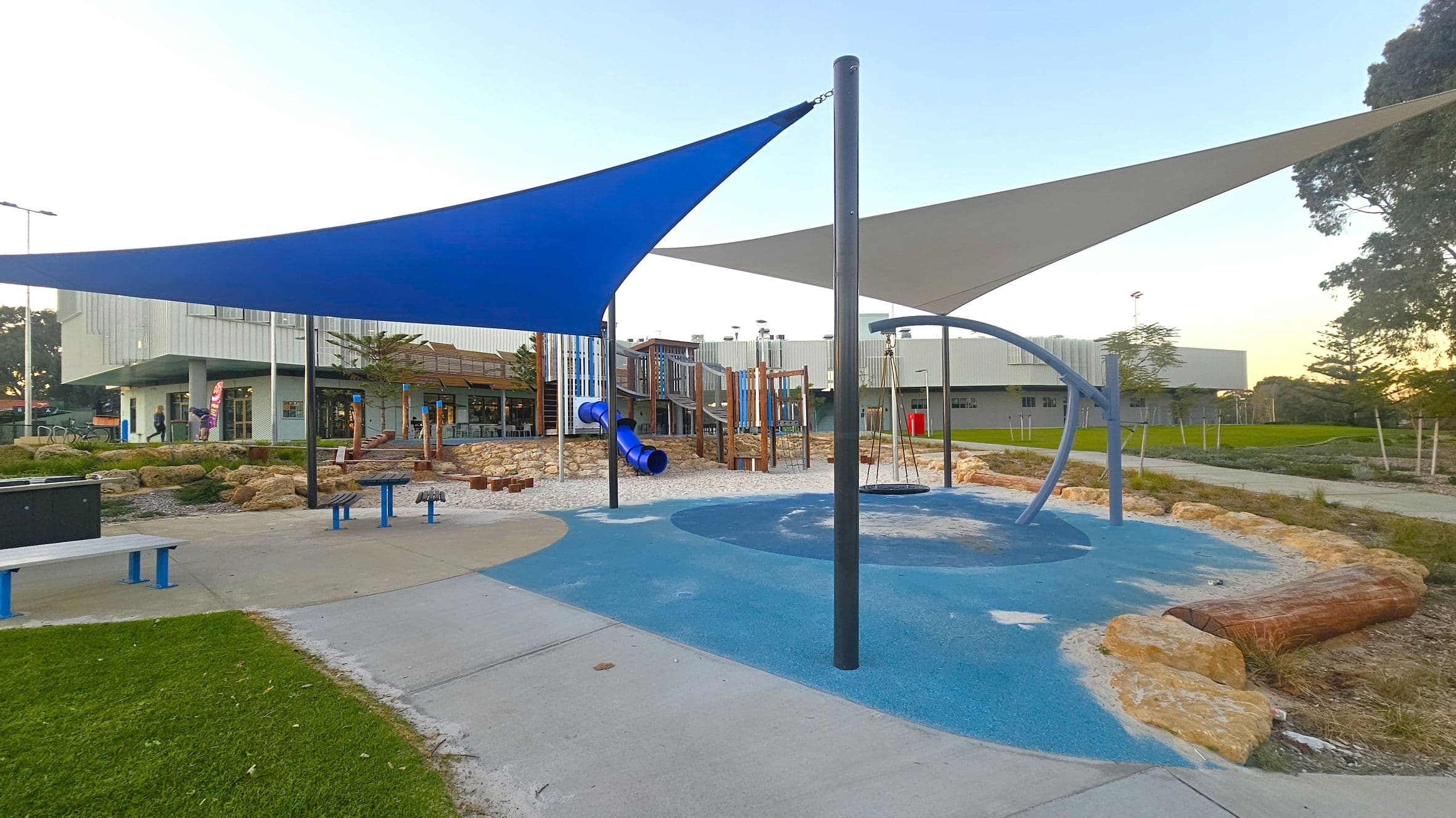 A playground with a blue and grey shade sail overhead. In the centre, there's a blue circular swing and a blue tube slide leading to a sandy area with wooden climbing structures. The ground is a mix of blue and grey soft play surfaces, with a concrete path and grass on the left. In the background, a modern building with large windows and a few people walking nearby. Trees and landscaped areas surround the playground.
