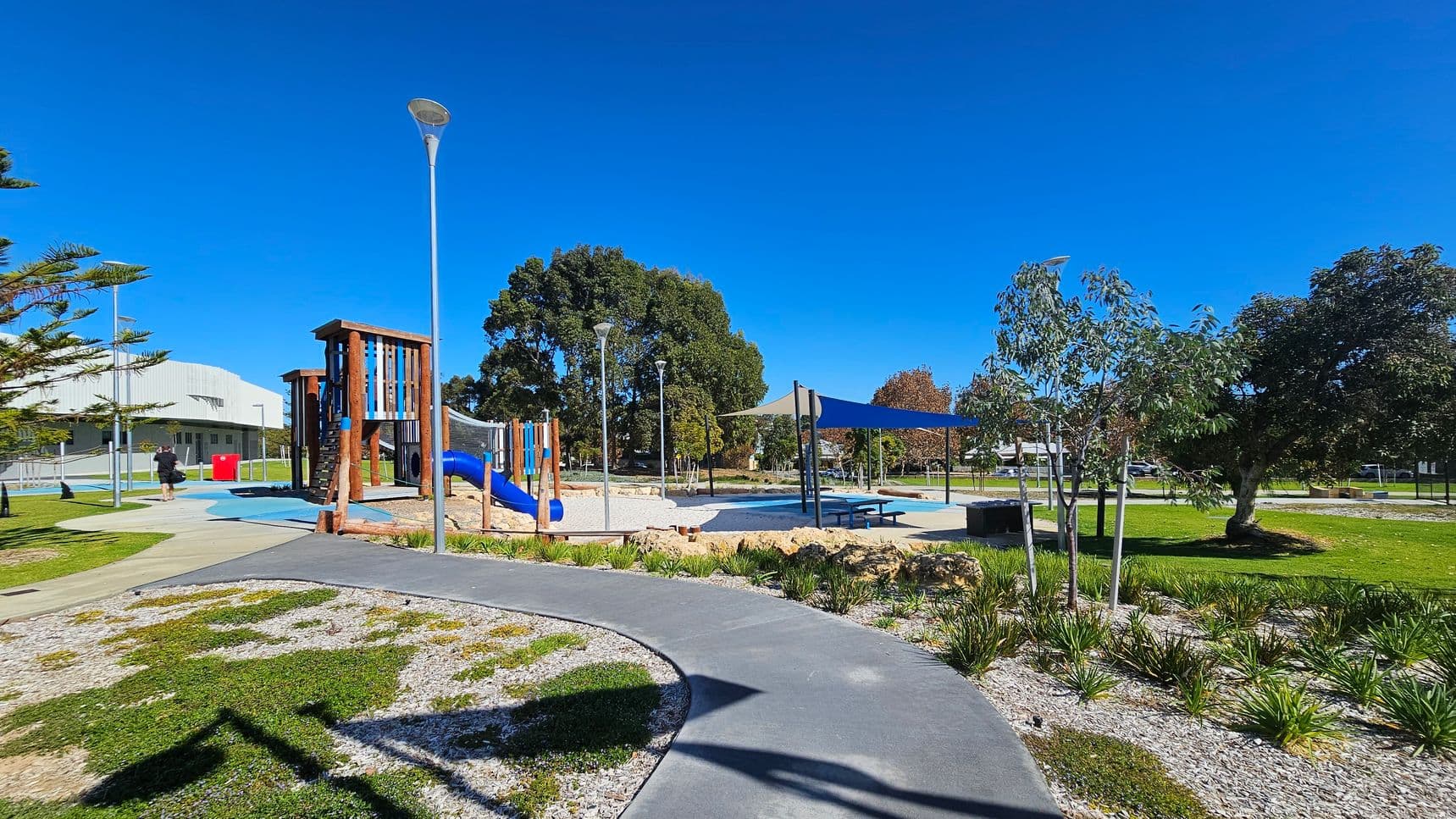 A playground with a wooden climbing structure and blue slide is on the left. A path curves through the foreground, bordered by grass and small plants. In the middle, a shaded area with a picnic table is covered by blue canopies. Trees are scattered throughout the park, with a large tree on the right. A building is visible in the background on the left, and several lamp posts are placed around the area.