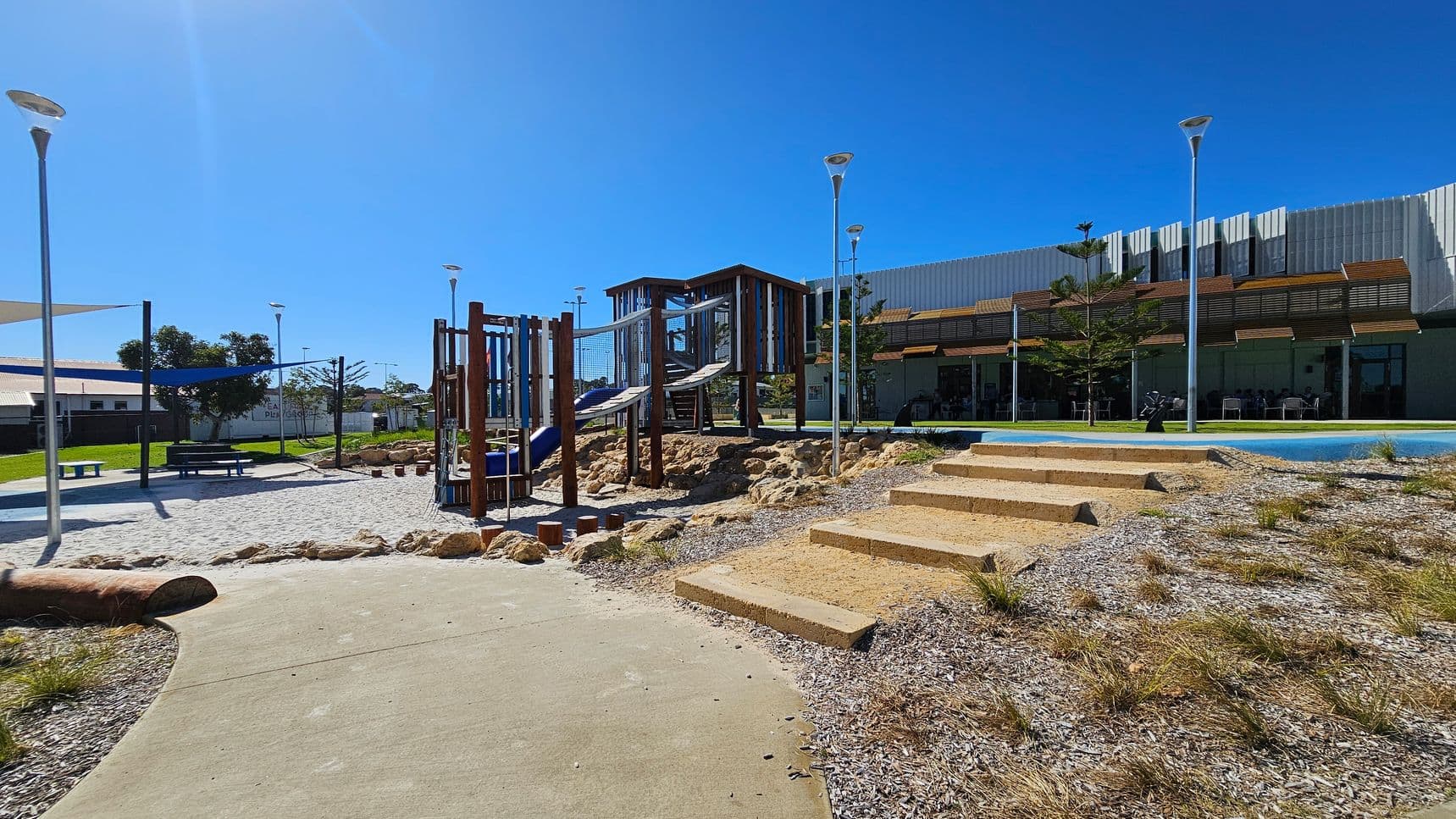 A playground with a climbing structure and slides is in the centre, surrounded by rocks and a sandy area. On the left, there are benches under shade sails and a grassy area with trees. On the right, a set of concrete steps leads up to a building with a modern design, featuring large windows and a shaded patio area. Several tall street lamps are scattered throughout the area. The ground is a mix of concrete paths and gravel with sparse grass.