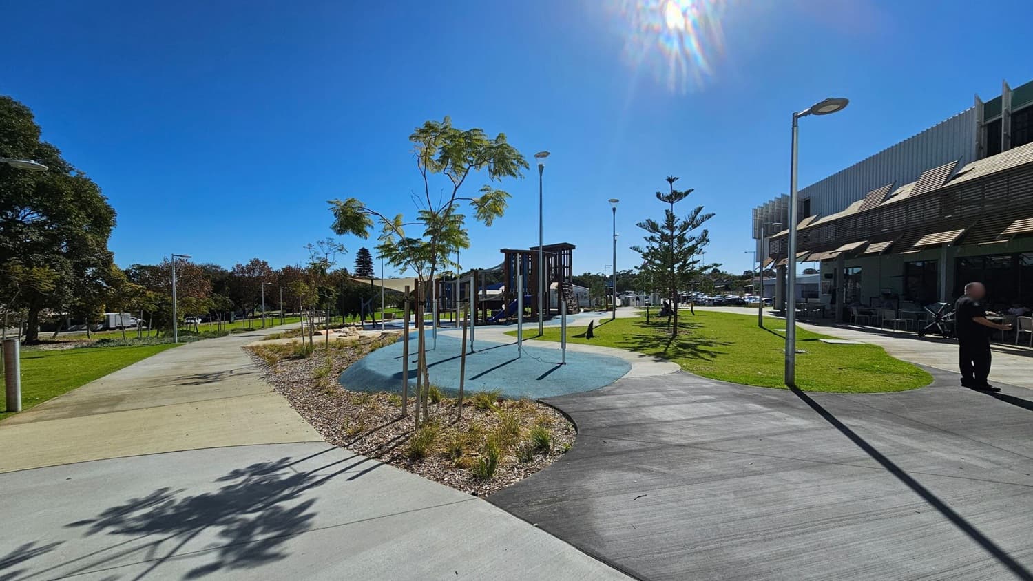 A park with a paved walkway curving through the centre, bordered by young trees and landscaped areas with mulch and grass. On the left, a larger tree and more greenery are visible. To the right, a modern building with outdoor seating and a person standing nearby. Several street lamps line the path, and a playground structure is visible in the background. The sky is clear and sunny.