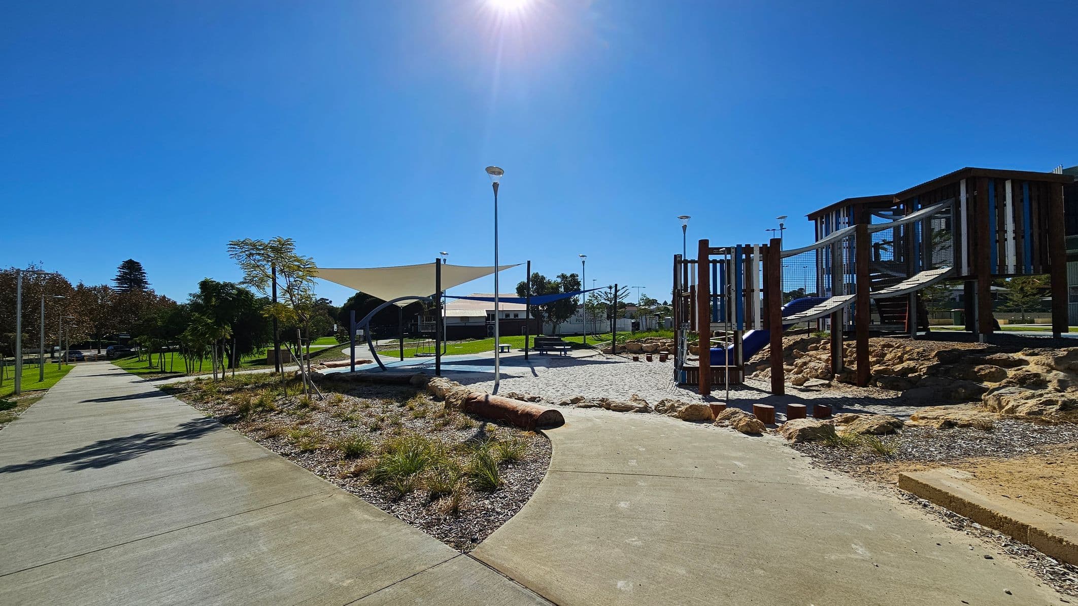 A playground with a modern design. On the right, there is a wooden play structure with slides and climbing nets, set on a rocky surface. In the centre, a shaded area with a sail-like canopy covers a seating area with a picnic table. On the left, a paved path runs alongside grass and small trees. The background features more greenery and a few buildings. The sky is clear and sunny.