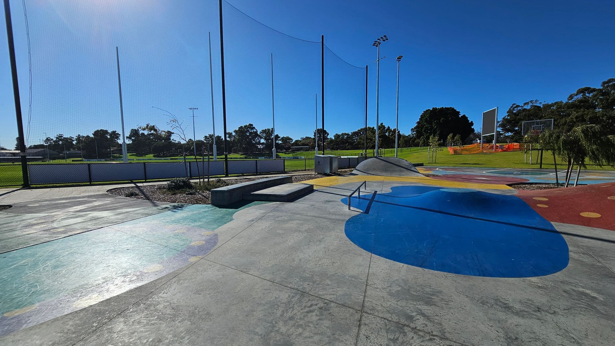 A skate park with a smooth concrete surface featuring colourful painted sections in blue, yellow, and red. In the centre, there is a small ramp and a rail for tricks. To the left, a low platform is visible. In the background, a grassy field is enclosed by a fence with tall poles and netting. Trees and a basketball hoop are on the right, with a large scoreboard nearby. The sky is clear and blue.