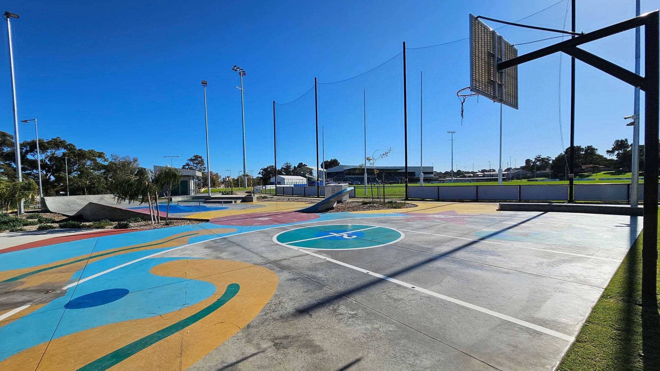 A colourful outdoor sports court with abstract patterns in blue, yellow, and orange on the ground. On the right, a basketball hoop with a damaged net is visible. In the background, there are tall poles with netting, a grassy field, and a building. Trees and shrubs are scattered around the area, and the sky is clear. The court is surrounded by a low fence.