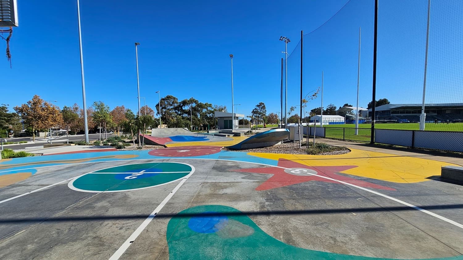 A colourful skate park with various ramps and painted designs on the concrete surface. In the background, there are tall light poles and a fenced sports field with green grass. Trees with autumn foliage line the left side, and a small building is visible in the centre. The sky is clear and blue.