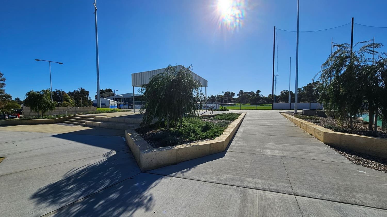 A paved area with a central path leading to a small building with a metal roof. On either side of the path are raised garden beds with small trees and shrubs. To the left, there are steps leading up to a grassy area with more trees and a few streetlights. In the background, a green field is visible, bordered by a fence and tall poles. The sun is bright in the sky, casting shadows on the ground.