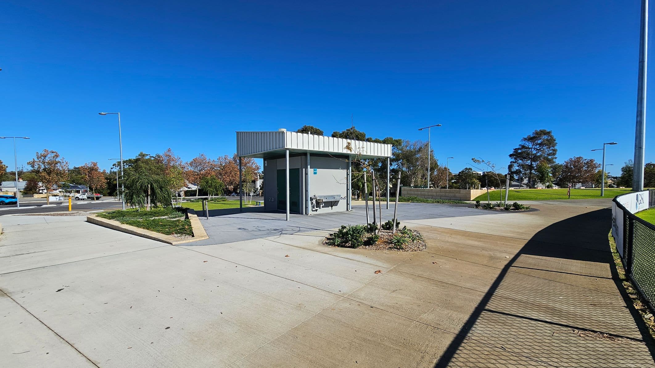 A small, modern public restroom building is centrally located, with a flat roof and grey walls. In front, there are a few small plants and a drinking fountain. The foreground features a concrete path with a shadow from a nearby fence on the right. To the left, a road with parked cars and street lamps is visible, bordered by trees with autumn foliage. The background includes a grassy area with more trees and additional street lamps.