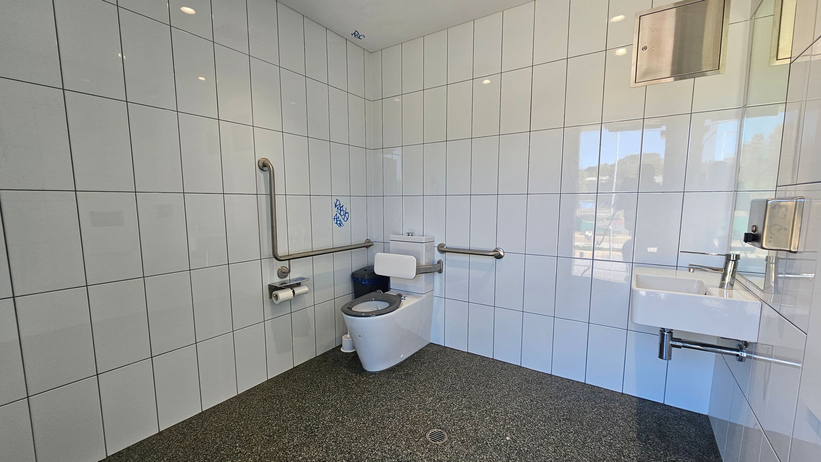 A public restroom with white tiled walls and a speckled dark grey floor. On the left, there are metal grab bars and a toilet with a black seat. Toilet paper rolls are mounted on the wall. In the centre, a small blue graffiti is visible above the toilet. On the right, a white sink with a metal tap is mounted on the wall, accompanied by a soap dispenser. A metal panel is above the sink. The ceiling is white with recessed lighting.