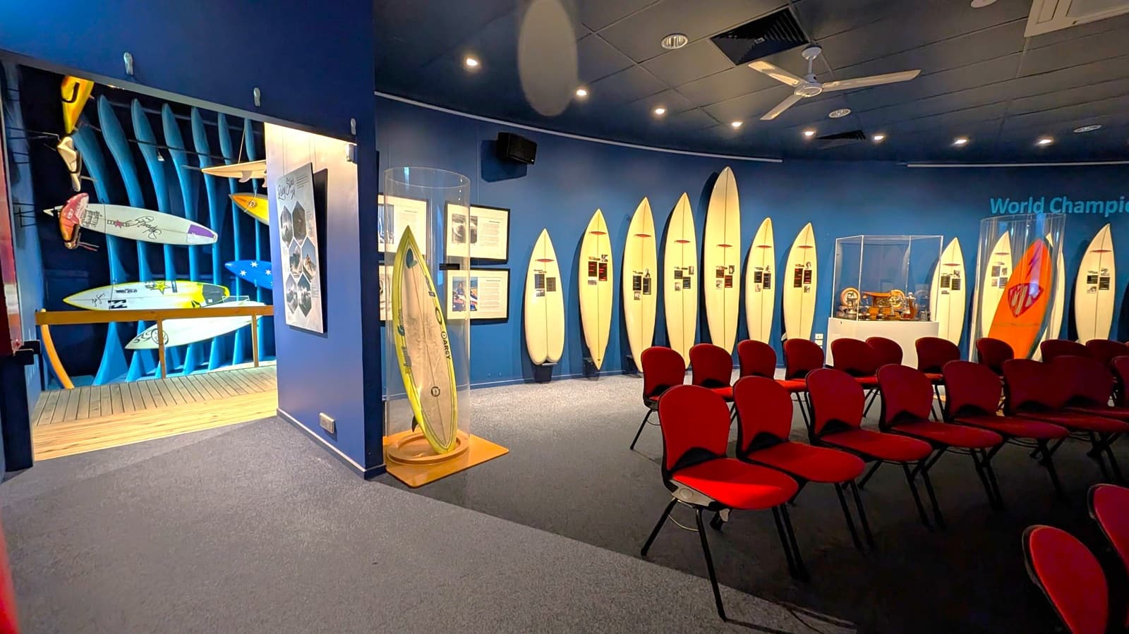 A museum exhibit with a blue floor and ceiling. On the left, there are display cases with various surfing equipment and wetsuits. The back wall features three surfboards in orange, blue, and yellow, mounted vertically. To the right, a large glass cabinet displays smaller surfing-related items. The walls are painted blue, and there are informational panels and photographs throughout the space.