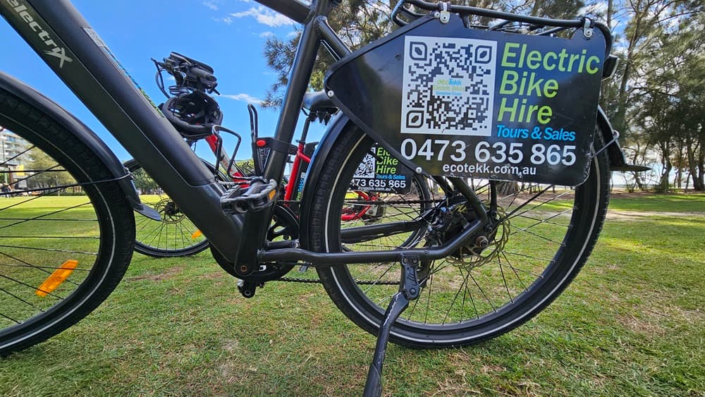 On the left, there is a close-up view of the rear wheel and frame of an electric bike. The middle features a QR code and a sign attached to the bike, displaying contact information for electric bike hire, including a phone number and website. On the right, another bike is partially visible. The background consists of a grassy park area with trees.