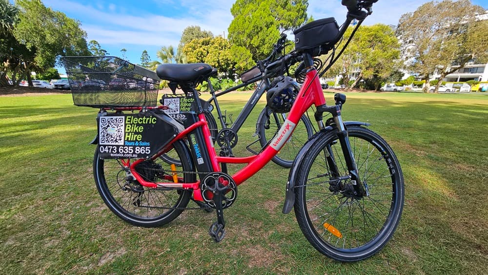 On the left, a red electric bike with a black basket on the rear and a sign displaying "Electric Bike Hire" along with a phone number and QR code. In the middle, the bike's frame and battery are visible. On the right, a grey electric bike is partially visible. The background consists of a grassy park area with trees and some parked cars.