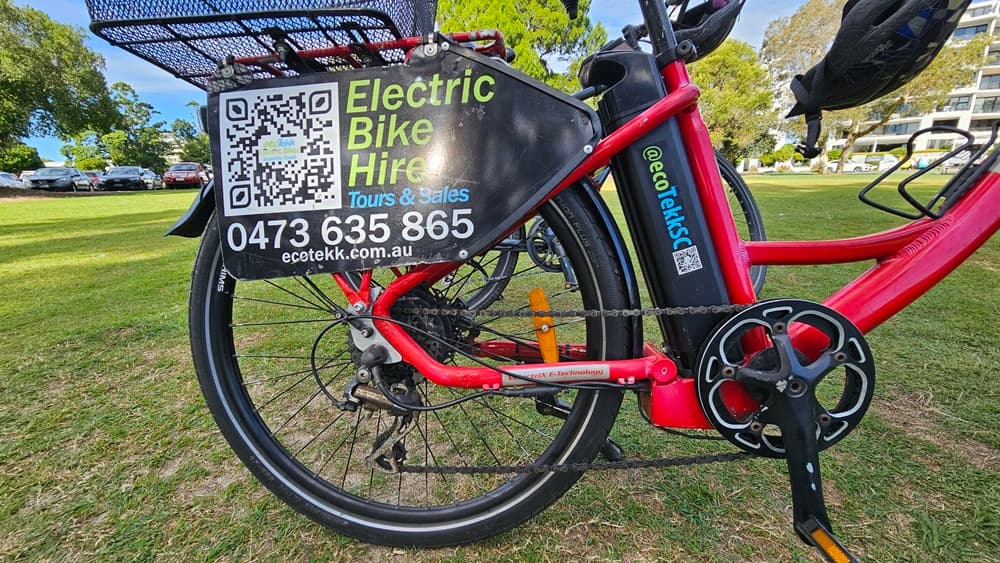 On the left, a red electric bike with a large black sign attached to the rear wheel, displaying "Electric Bike Hire," a QR code, a phone number, and a website. In the middle, the bike's frame, battery, and part of the pedal are visible. On the right, another red electric bike is partially visible. The background consists of a grassy park area with trees and parked cars.