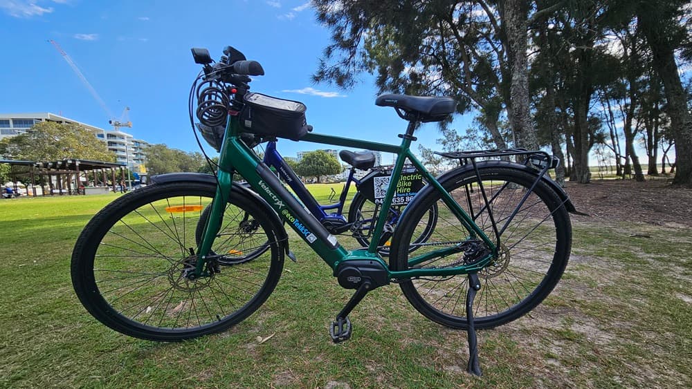 On the left, a dark blue electric bike with a small black bag attached to the top tube. On the right, a green electric bike with a similar design, featuring a black rear rack. In the middle, the green bike's frame and pedals are clearly visible. The background includes a grassy park area with trees, a pavilion, and a distant building.