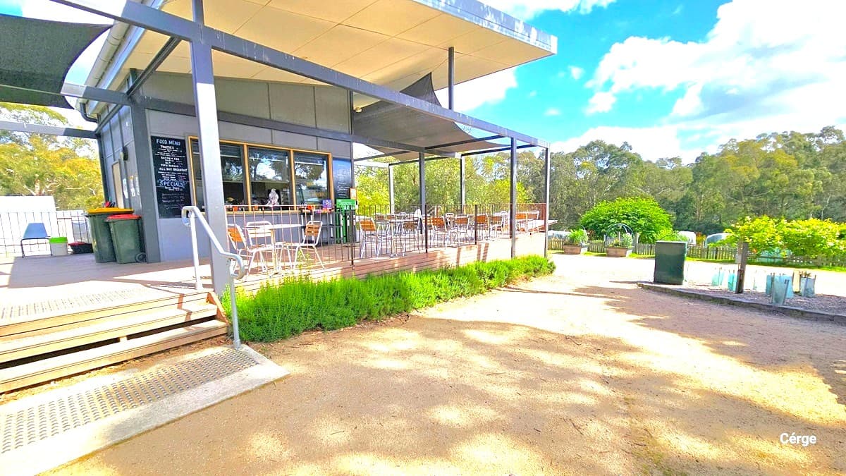 Entrance to Cafe Eden. The cafe is painted with a grey and white ceiling. A menu is written on black board placed beside the glass window. Tables and chairs are arranged on the deck. On the left, there is a two-tier staircase with a handrail. Two green rubbish bins are placed against the wall, and a plant box with green shrubs is in front. To the right, a shade cloth is also placed overhead.
