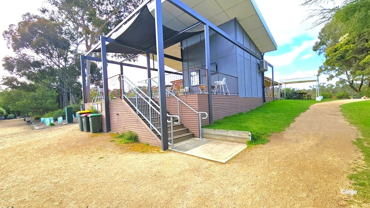 The cafe is constructed with timber and steel, painted in grey and white. Another entrance to the cafe is through a staircase with a handrail. Truncated domes are installed before and after the stairs for safety. Tables and chairs are arranged on the deck. On the left, rubbish bins are placed against the wall. To the right, a dirt path between a grassy area leads to a shaded area