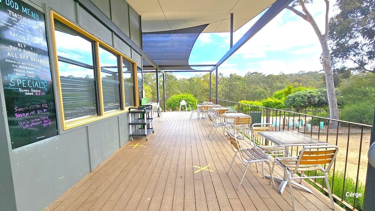 Outside the cafe, the food menu is displayed on a blackboard affixed to the grey wall beside the glass window. A food trolley stands against the wall. Neatly lined up on the right are tables and chairs. Overhead, a black shade cloth provides shade to the tables. The wooden floor is marked with yellow "X" markings. Trees and shrubs surround the area.