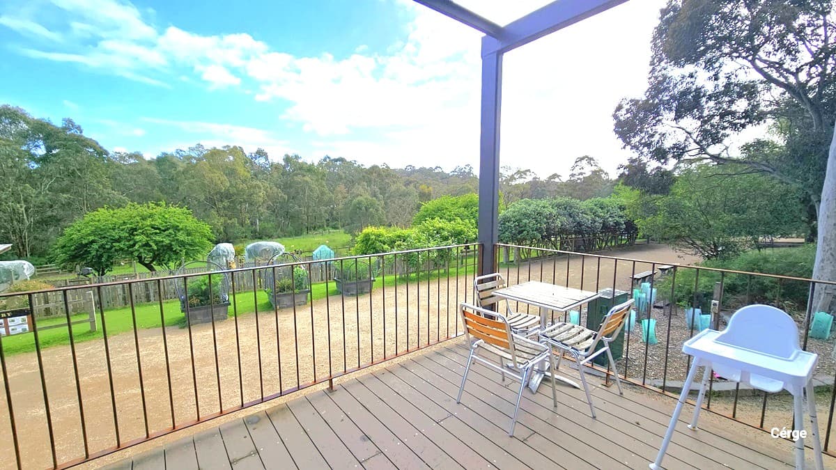 In the corner is a table with three chairs, accompanied by a white high chair for babies. Neatly lined up on the right are tables and chairs, with a wooden floor. Black metal railings secure the area, and small trees in green plastic fencing adorn the ground. Larger trees and green grass surround the area.