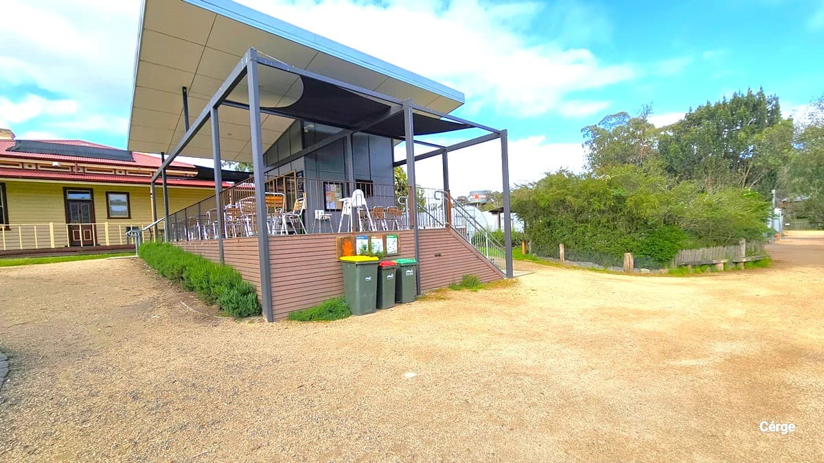 Another view of the cafe reveals a structure made of timber and metal, painted in cream and grey. The lower wall exhibits a corrugated design. Three rubbish bins and poster signages are strategically placed. To the right, there's an ornamental landscape extending towards another entrance. The white-ceilinged structure features additional stairs serving as an entrance on the right. Trees and bushes surround the area.