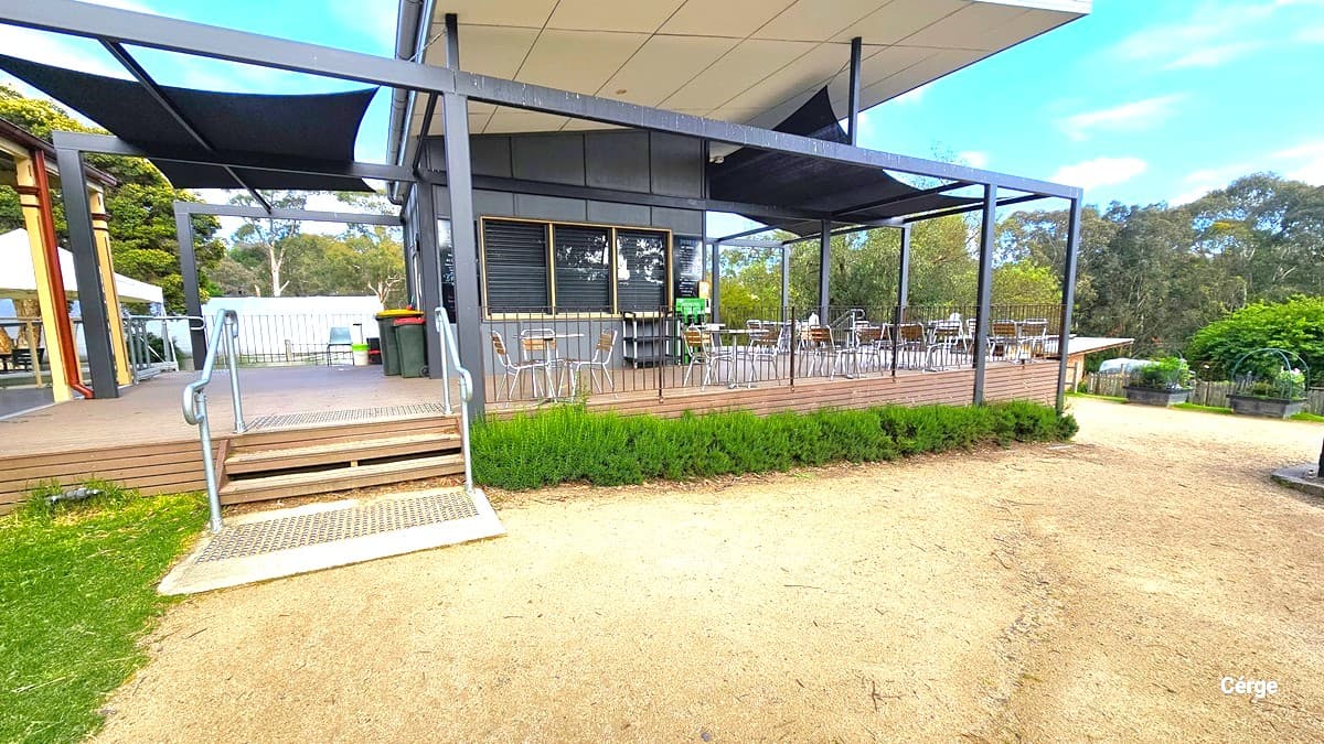 The entrance to Cafe Eden reveals a grey and white-painted structure. A menu on a blackboard is positioned near the glass window. Tables and chairs are neatly arranged on the deck. On the left, a two-tier staircase with a handrail is present. Two green rubbish bins are against the wall, accompanied by a plant box with green shrubs in front. To the right, shade cloth is also placed on both sides.