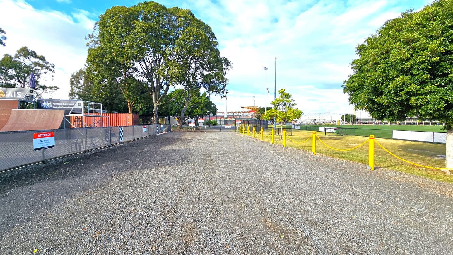 A gravel path runs through the centre, flanked by trees on both sides. On the left, there's a fenced area with a skate ramp and a sign that reads "Attention No entry." On the right, a grassy field is bordered by a yellow rope barrier. In the background, there are more trees and a sports field with floodlights. The sky is partly cloudy.