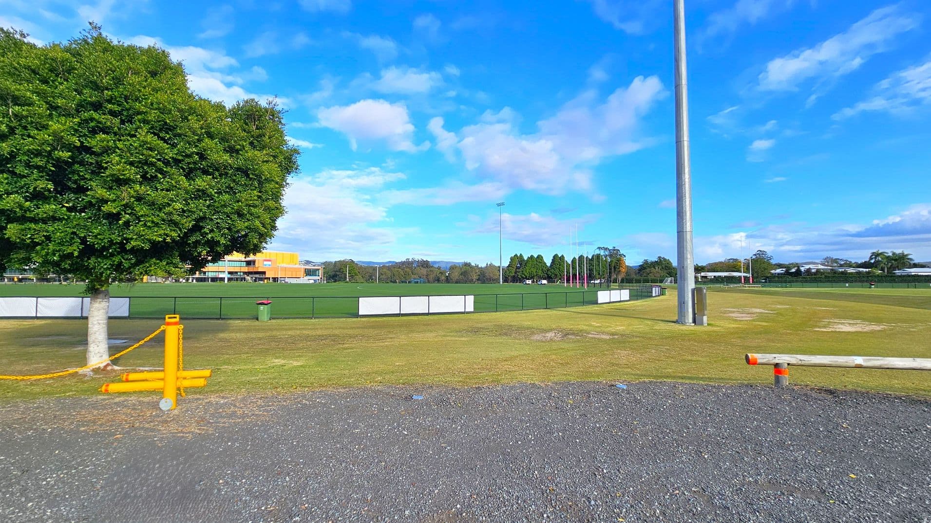 A grassy sports field with a tall floodlight pole on the right. In the foreground, there's a gravel path and a wooden barrier with orange markings. On the left, a large leafy tree stands next to a yellow chain barrier. In the background, a building with orange and yellow accents is visible, along with a line of trees and distant hills. A green rubbish bin is near the fence surrounding the field.