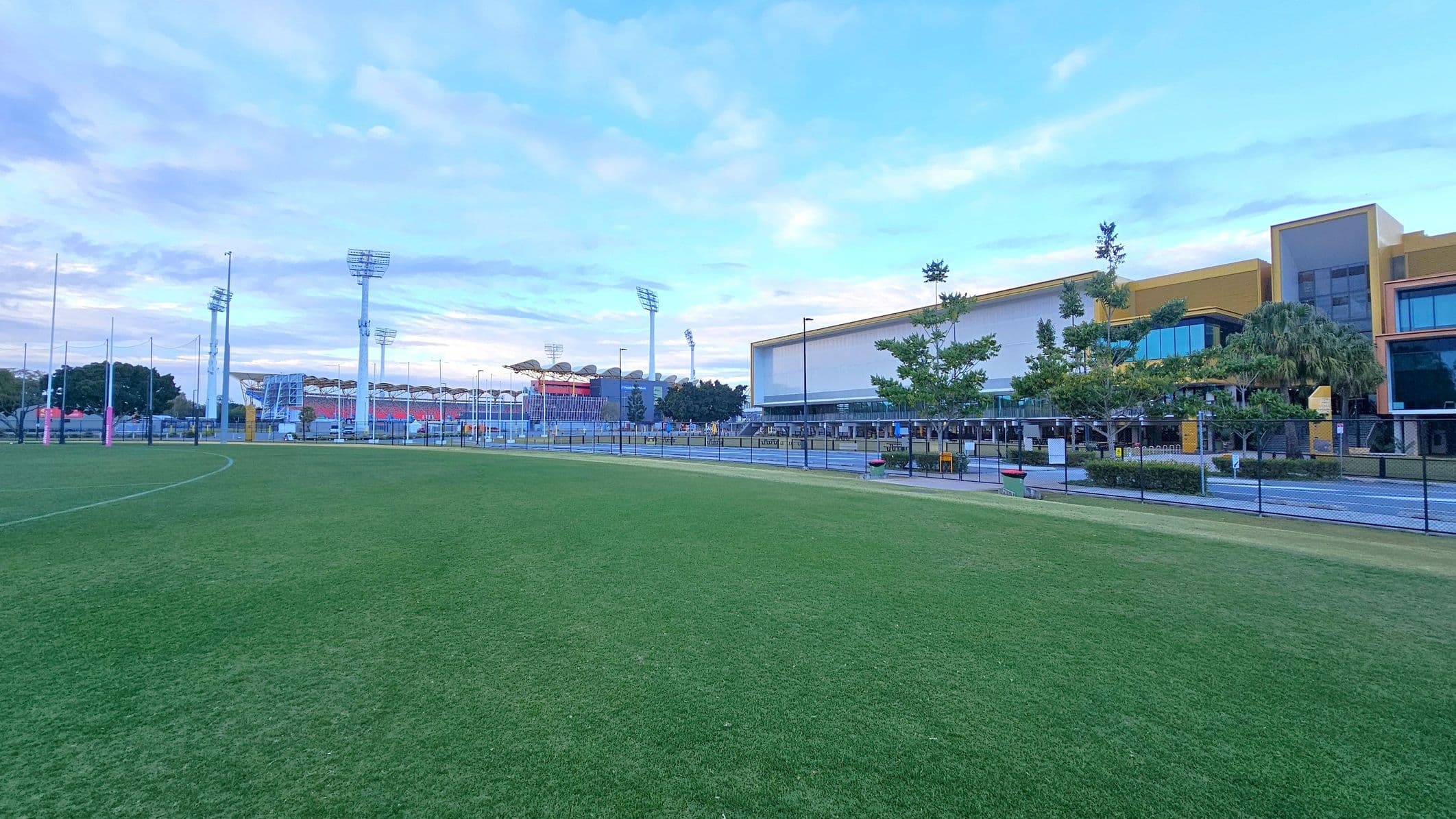 A grassy field in the foreground with a sports stadium in the background. The stadium has tall floodlights and a seating area with a red and white colour scheme. On the right, there is a modern building with large windows and a yellow and grey facade, surrounded by trees and a fence. The sky is partly cloudy.